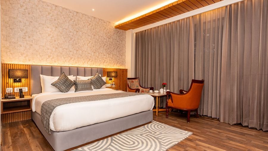 An image of a Deluxe Room with a bed, bed side table, lamps, rug and chairs and table - The Belvedere Himalayan Retreat, McLeodganj