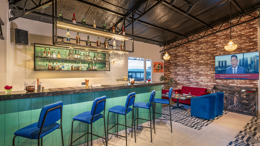 A rooftop bar with a teal counter, blue bar stools, and a well-stocked liquor shelf, a high ceiling with exposed beams, a brick accent wall, and a lounge area featuring blue and red seating at Lords Eco Inn Dehradun.