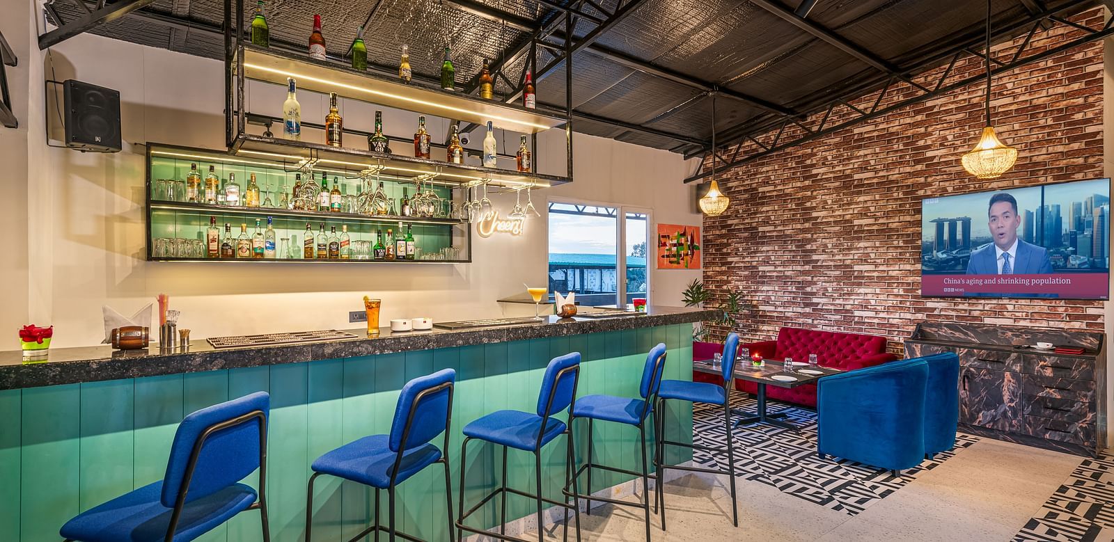 A bar with a teal counter, blue bar stools, and a well-stocked liquor shelf. It boasts a high ceiling with exposed beams, a brick accent wall, and a lounge area featuring blue seating - Lords Eco Inn Dehradun.