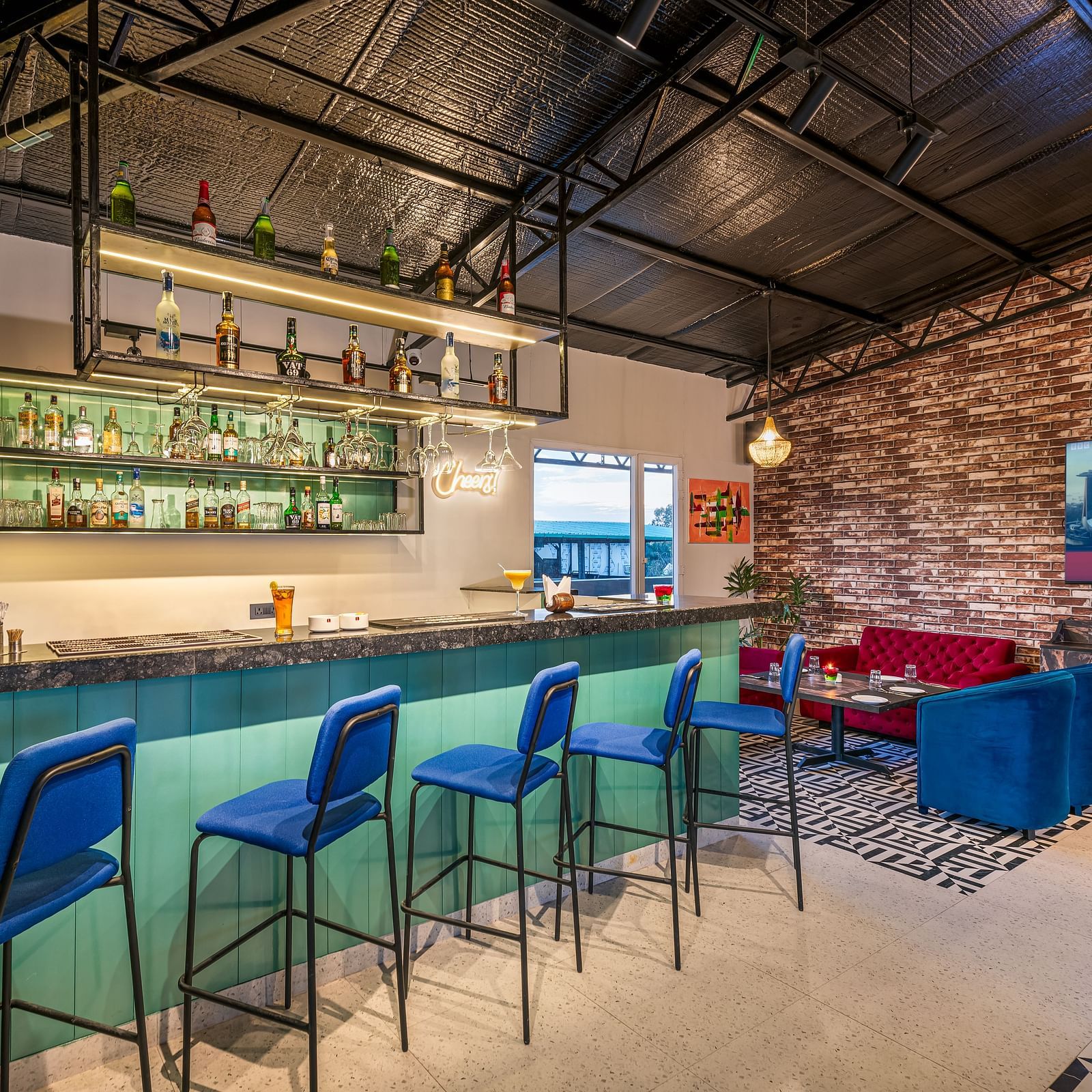 A bar with a teal counter, blue bar stools, and a well-stocked liquor shelf. It boasts a high ceiling with exposed beams, a brick accent wall, and a lounge area featuring blue seating - Lords Eco Inn Dehradun.