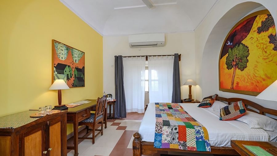 A bedroom with a bed, artwork, a ceiling fan, a desk, and arched architectural details at Tijara Fort-Palace - 19th Century, Alwar.