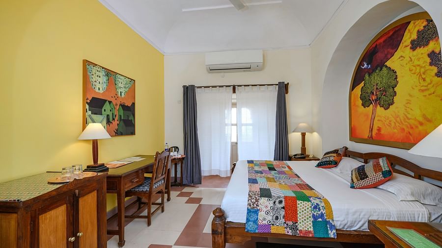 Bedroom with a bed, wooden furniture including desks and bedside tables with lamps, artwork, and an air conditioner at Tijara Fort-Palace, Alwar.