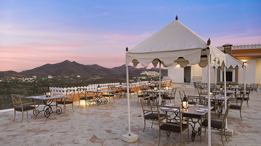 Shot of an outdoor dining area at Khas Mahal by Fateh Collection, Udaipur with white canopies and seating set against a backdrop of hills and a twilight sky.