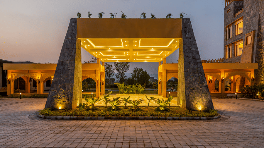 A lit stone portico and entryway at EsthereaRaj Leela, Ranakpur with a paved driveway and small plants along the base.