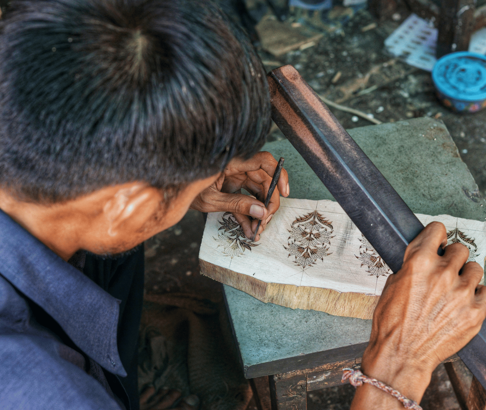 An artisan engraves intricate patterns onto a small piece of wood or paper using a long, dark metal tool.