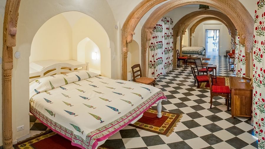 Room with bed, patterned bedding, arched doorways, patterned floor, furniture including chairs and tables, and patterned rugs - Tijara Fort-Palace - 19th Century, Alwar.