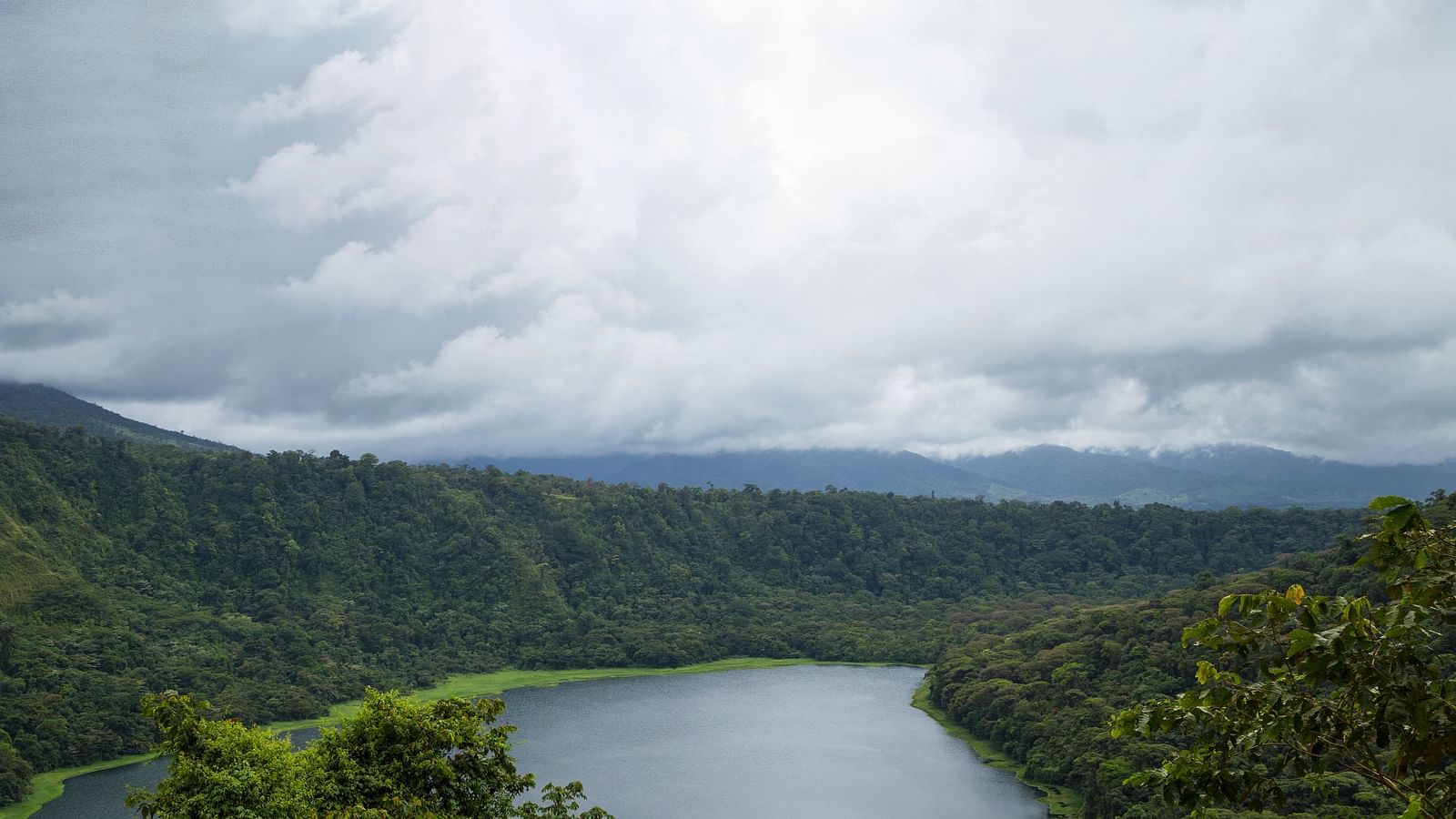 A peaceful lake nestled amidst forested hills with a cloudy grey sky.