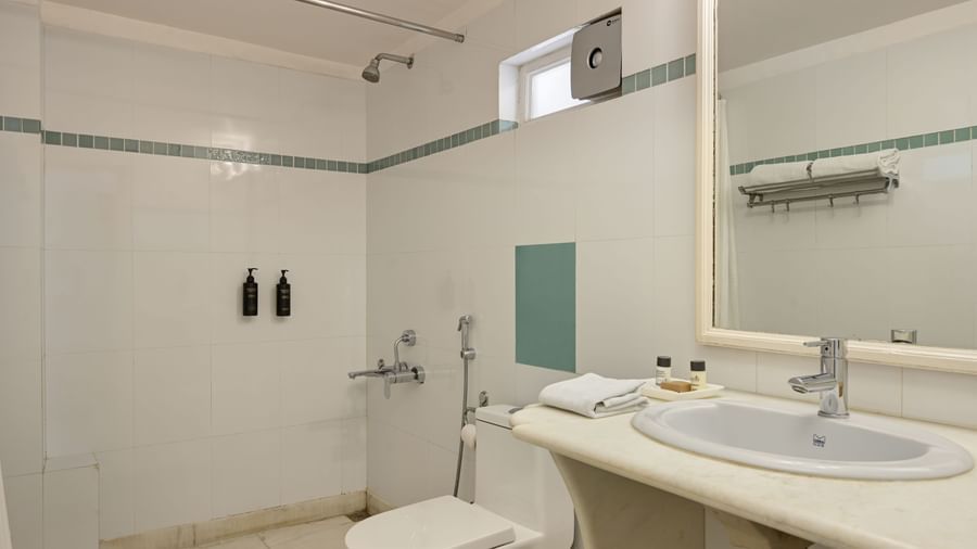 A white tiled bathroom at Ram Pratap Palace, Udaipur, featuring a large mirror, a sink with basic toiletries, a western toilet, and a shower area with a curtain rail.
