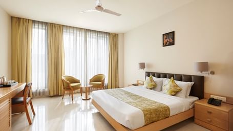 A well-lit hotel room at Monday Hotels Mahape, Navi Mumbai, with a large bed, seating area, and wooden furniture.