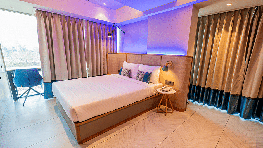  A spacious room at Indie Stays Mumbai International Airport features a large bed, windows on two sides, curtains fully drawn on one side, partially open on the other, and cosy seating by the window.
