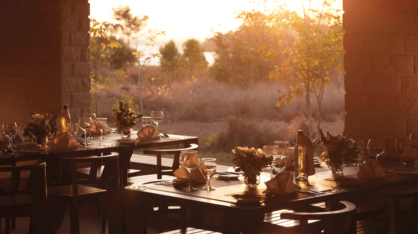 Interior view of Wildgrass restaurant at The Serai Kabini showing dining tables set with plates, glasses, and cutlery, facing an open wall with trees and sunlight visible outside.