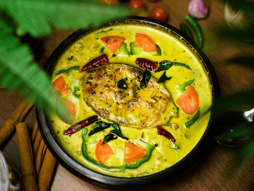 A top-down view of a bowl of fish curry with a piece of fish in the centre and slices of capsicum and tomatoes next to it.
