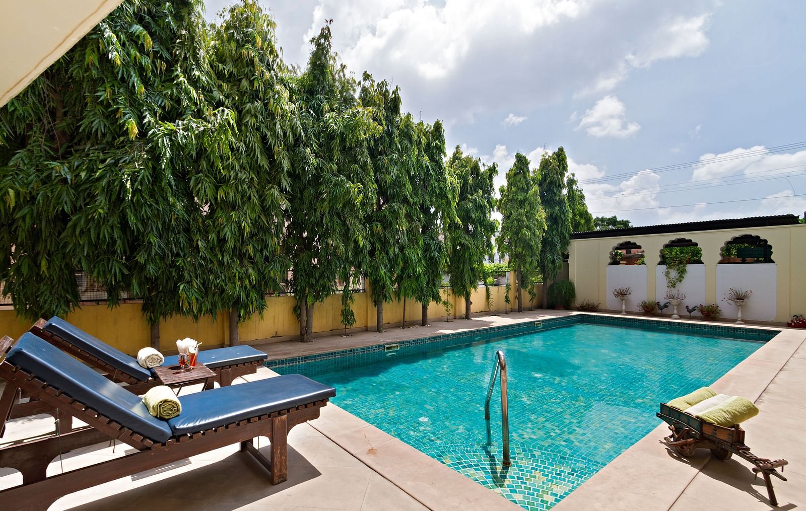 An outdoor swimming pool with blue water, surrounded by sun loungers and tall green trees, under a cloudy sky at Dera Rawatsar, Jaipur