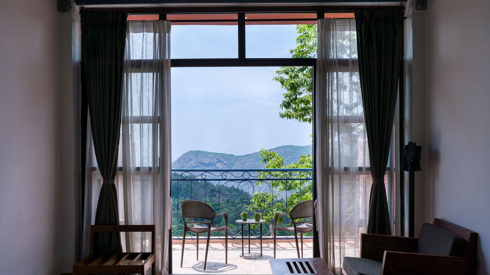 Room with a large window and glass doors leading to a balcony with a scenic mountain view, furnished with a cozy seating area. | Mango Hill, Yercaud