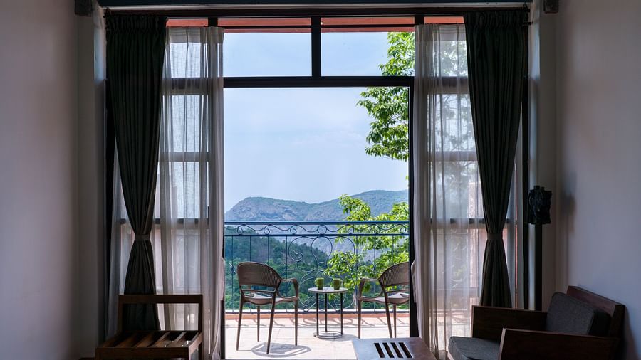 Room with a large window and glass doors leading to a balcony with a scenic mountain view, furnished with a cozy seating area. | Mango Hill, Yercaud