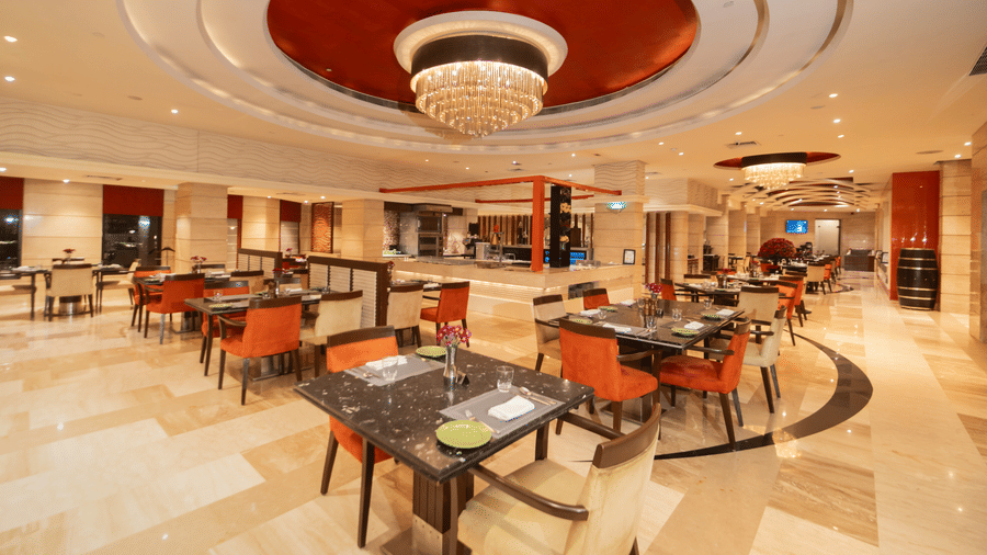 Ssence, the all-day dining restaurant at The Suryaa New Delhi, featuring a grand gold chandelier and elegantly arranged dining tables and chairs