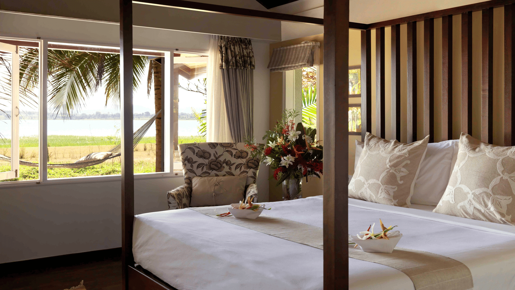 Bright Waterfront Villa bedroom at The Serai Kabini with a four-poster bed, river-facing windows, an armchair, floral accents, and serene views of nature for a relaxing stay in Kabini.