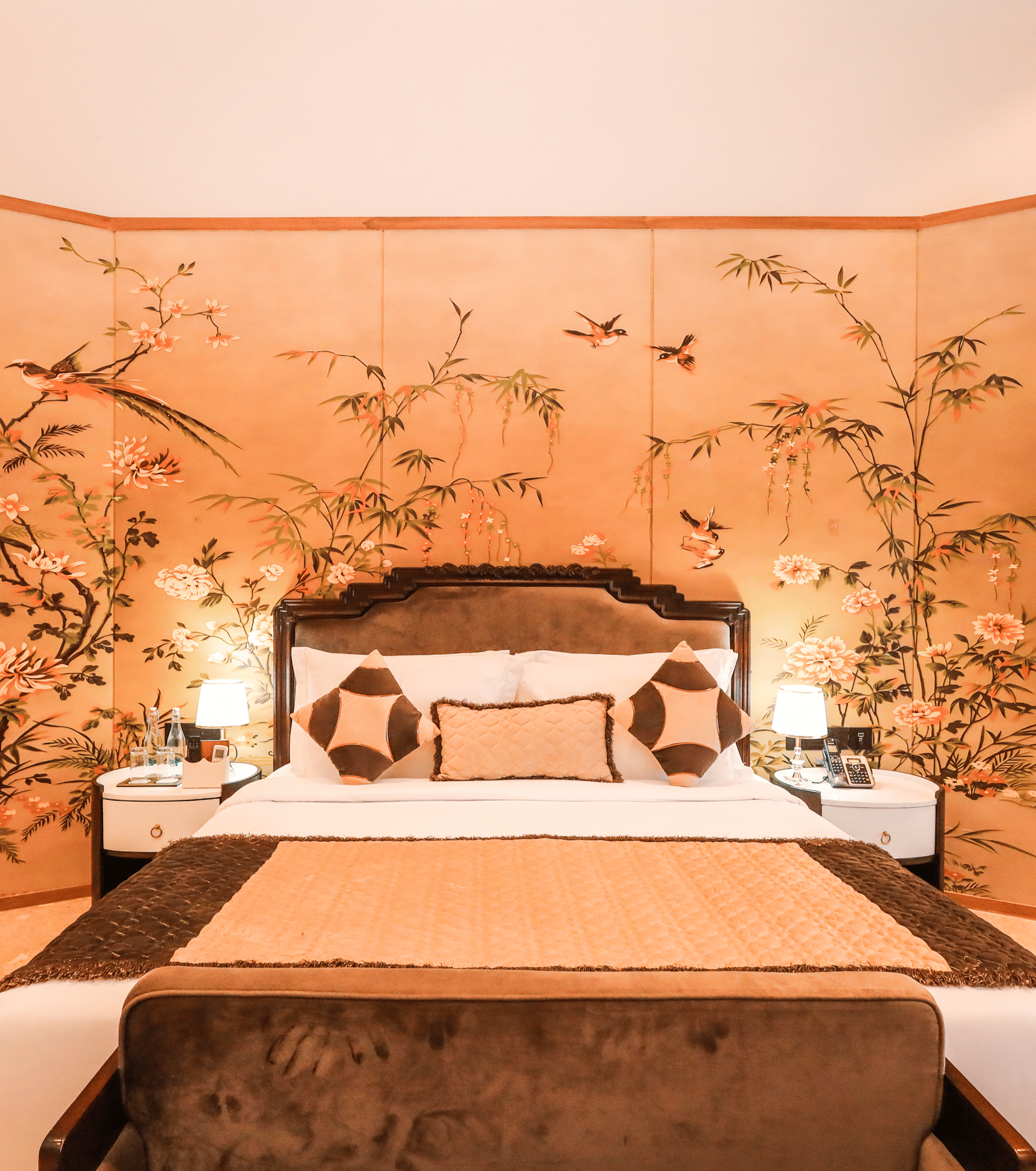An interior shows a bed with side tables, plants on both sides, a bench at the foot of the bed and a wall panel behind the bed at Ananta Spa & Resort, Jaipur.