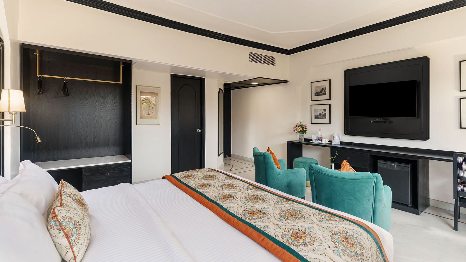 a king bed in a spacious hotel room in udaipur featuring two green armchairs, a TV, a wardrobe and several wall paintings - Hotel Rajdarshan, Udaipur.