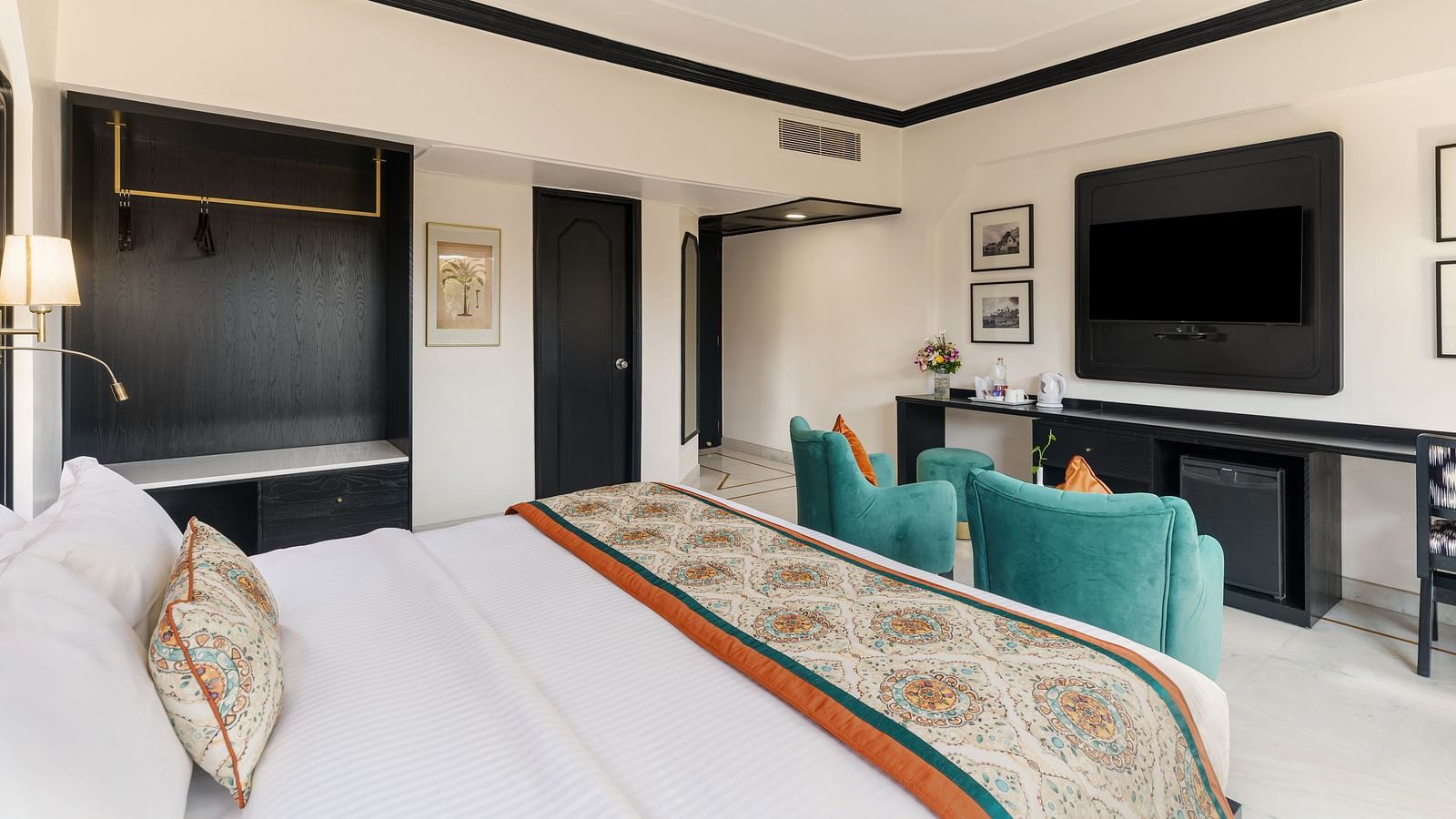 a king bed in a spacious room featuring two green armchairs, a TV, a wardrobe and several wall paintings - Hotel Rajdarshan, Udaipur.
