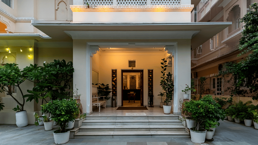 Steps leading to a lit building entrance flanked by potted green plants and a stone-paved courtyard at Akshay Niwas Boutique Hotel by Amantra, Udaipur.