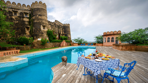  Exterior view of Hill Fort-Kesroli, featuring a table and chair facing a swimming pool under a cloudy sky.