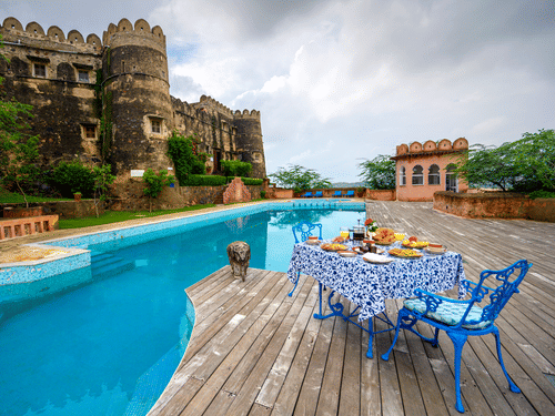  Exterior view of Hill Fort-Kesroli, featuring a table and chair facing a swimming pool under a cloudy sky.