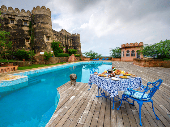  Exterior view of Hill Fort-Kesroli, featuring a table and chair facing a swimming pool under a cloudy sky.