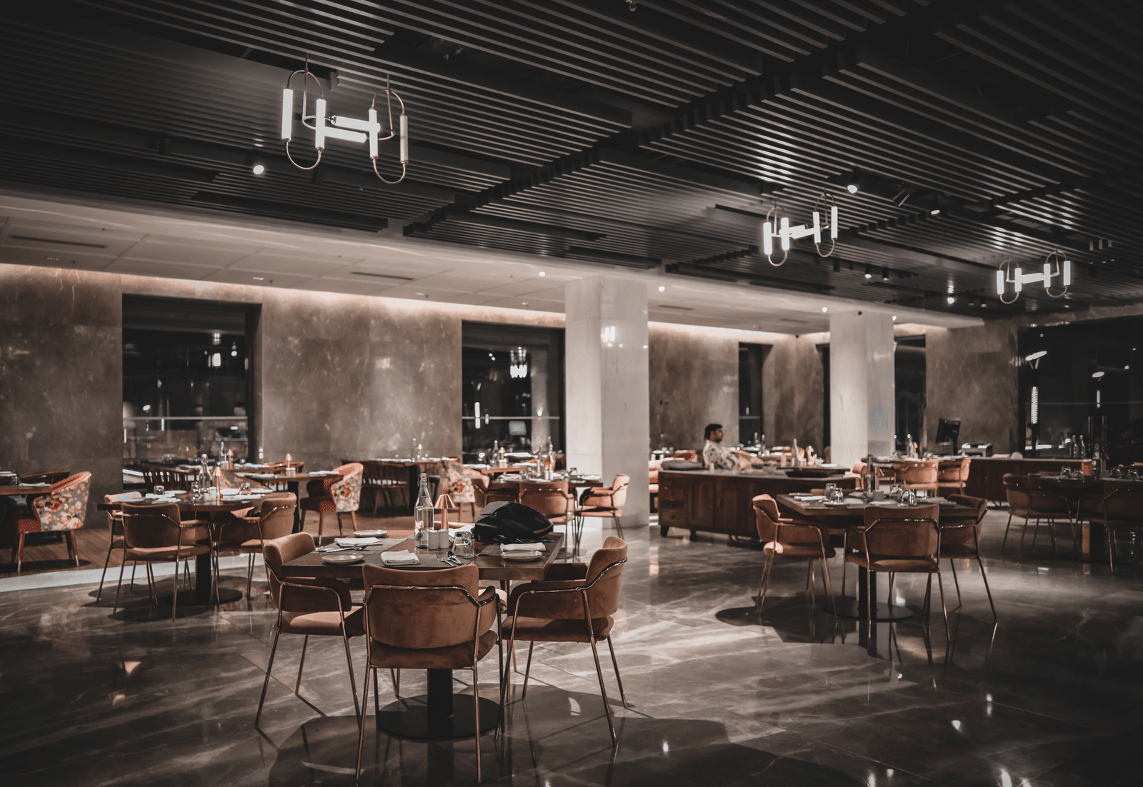A wide-angle shot of a modern restaurant dining room with empty tables and chairs. The room has warm lighting and large windows.