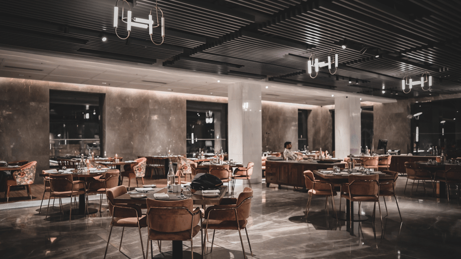 A wide-angle shot of a modern restaurant dining room with empty tables and chairs. The room has warm lighting and large windows.