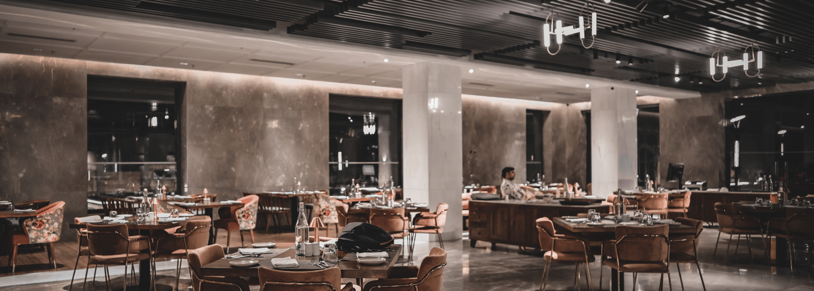 A wide-angle shot of a modern restaurant dining room with empty tables and chairs. The room has warm lighting and large windows.