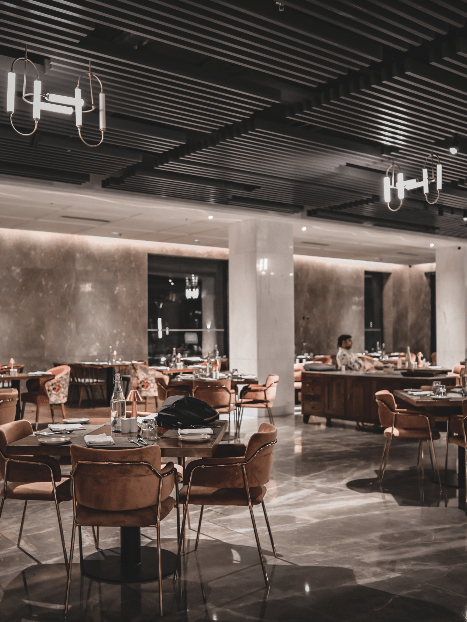 A wide-angle shot of a modern restaurant dining room with empty tables and chairs. The room has warm lighting and large windows.