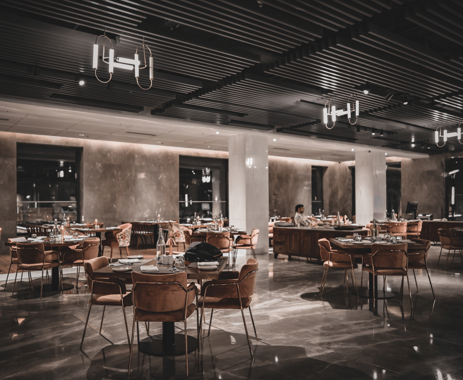 A wide-angle shot of a modern restaurant dining room with empty tables and chairs. The room has warm lighting and large windows.