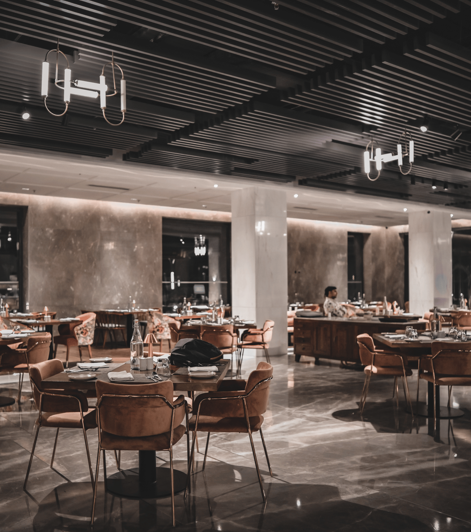 A wide-angle shot of a modern restaurant dining room with empty tables and chairs. The room has warm lighting and large windows.