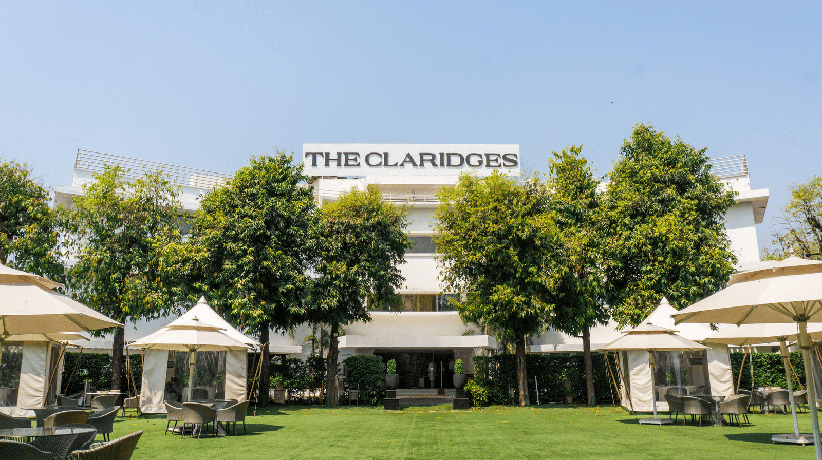 Exterior view of The Claridges hotel building featuring a manicured green lawn, surrounding greenery, and outdoor umbrella seating.