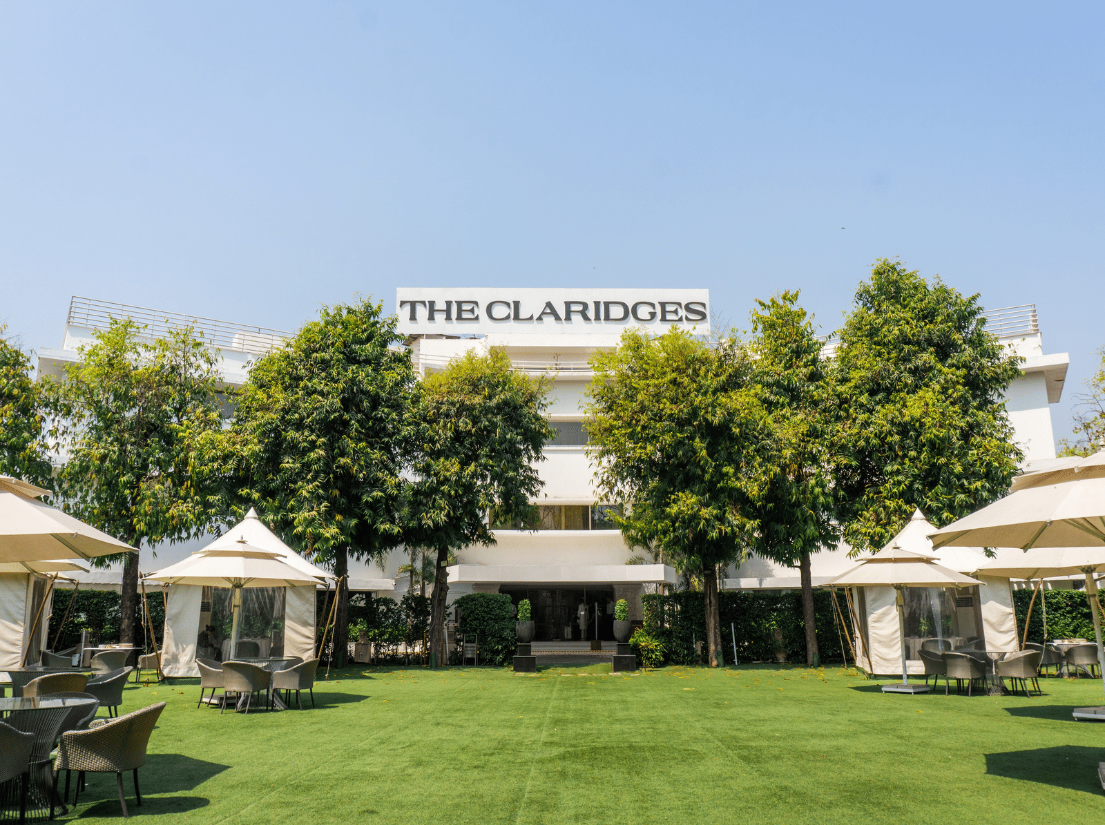 Exterior view of The Claridges hotel building featuring a manicured green lawn, surrounding greenery, and outdoor umbrella seating.