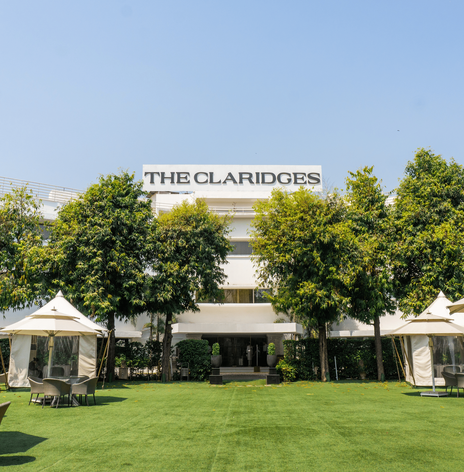 Exterior view of The Claridges hotel building featuring a manicured green lawn, surrounding greenery, and outdoor umbrella seating.