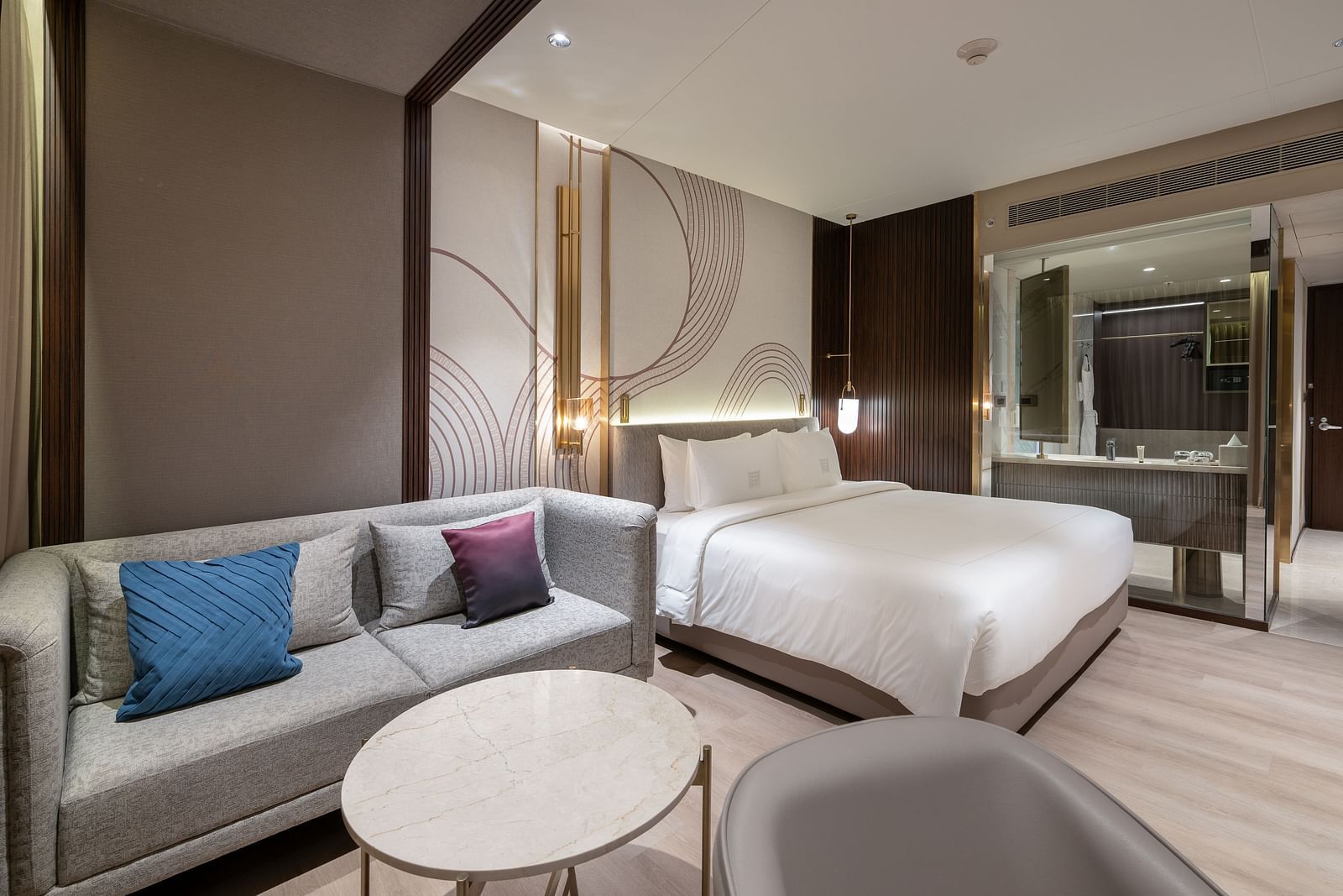 A modern hotel room with a large bed, a comfortable grey sofa, and a round coffee table at Merlis Hotel, Coimbatore.