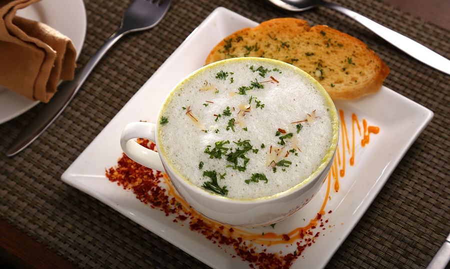 A cup of creamy mushroom soup, garnished with herbs and saffron, served on a square plate with toasted bread, orange sauce swirl, and red spice. A fork, spoon, and napkin rest on the brown placemat beside it at Uday Samudra Leisure Beach Hotel.