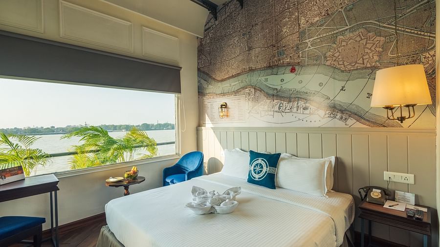Room with a view of a river through a large window, featuring a wall map, queen bed, and blue armchair.