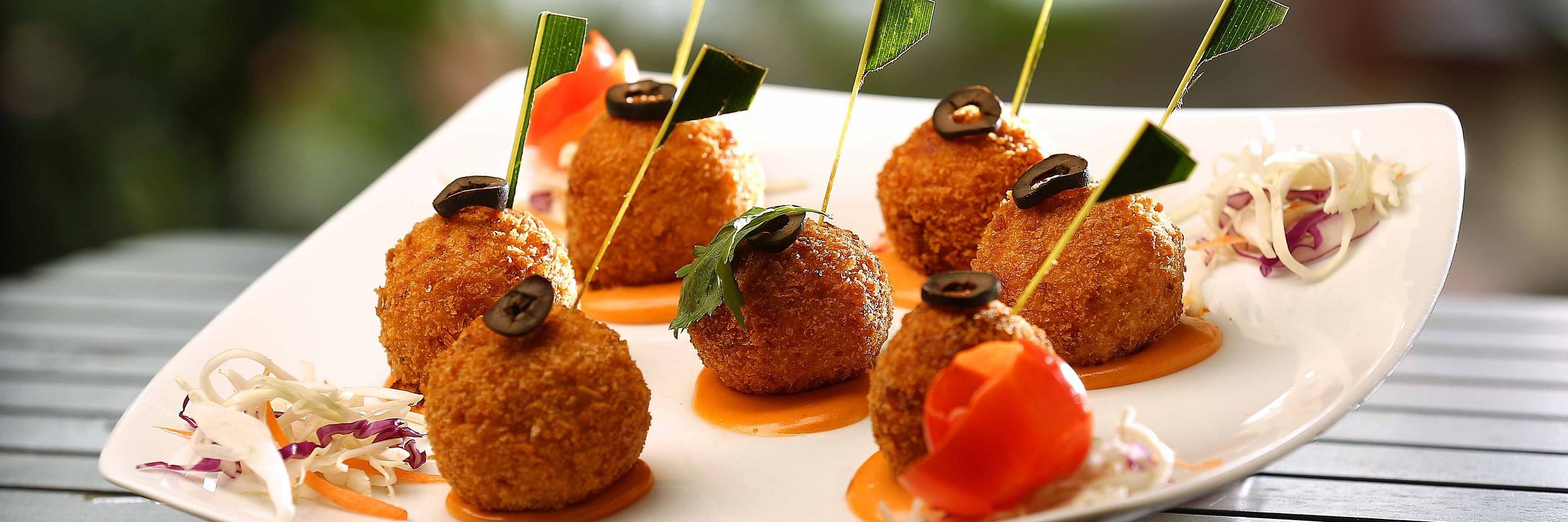 A white plate on a striped surface holds six golden-brown, round appetisers, each topped with a black olive and a small flag. They are arranged with dollops of white and orange sauce, shredded carrots, and cabbage at Uday Samudra Leisure Beach Hotel.