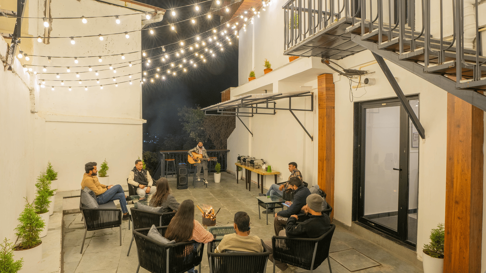 A group of people gathered in the common area of the Perfectstayz Premium Westend Mall Road, Mussoorie, decorated with fairy lights during the late evening hours.