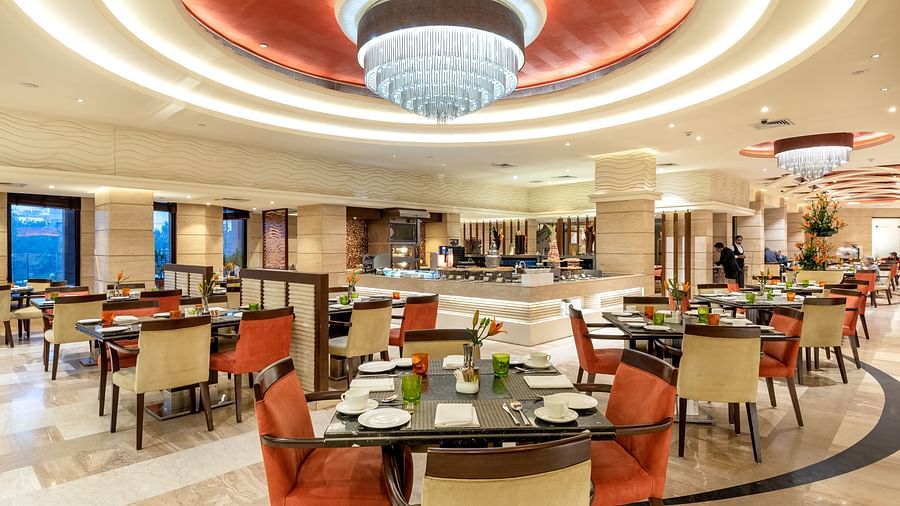 An image of a well lit dining hall where several sets of dining table and chairs are placed - The Suryaa New Delhi