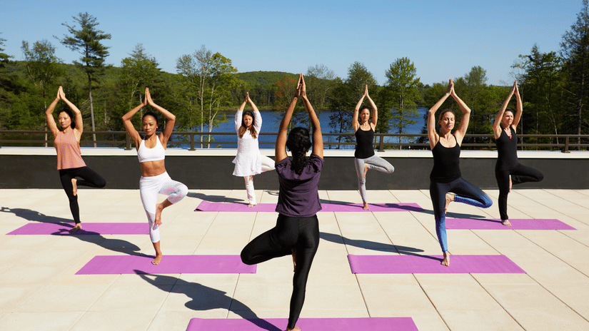 A group of 7 women performing yoga on the rooftop at YO1 Longevity & Health Resorts, Catskills with a backdrop of the lake surrounded by trees