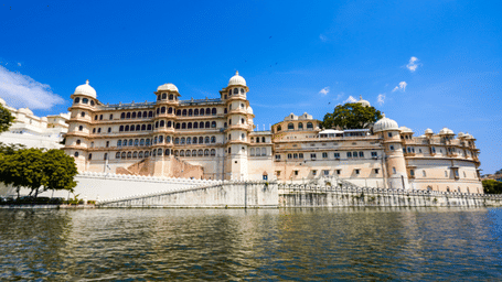 Wide-angle view of the City Palace in Udaipur, Rajasthan, overlooking Lake Pichola, featuring grand white and beige architecture with domes, balconies, and reflections in the water under a clear blue sky.