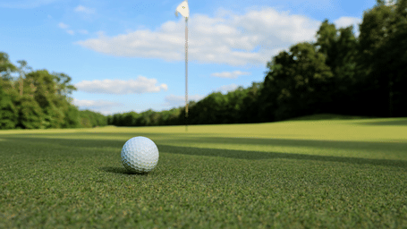Close-up of a white golf ball on a manicured green with a distant flagstick and tree line under a blue sky with light clouds.