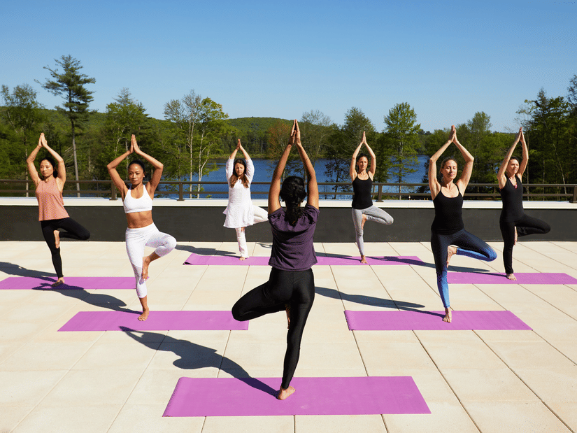 A group of 7 women performing yoga on the rooftop at YO1 Longevity & Health Resorts, Catskills with a backdrop of the lake surrounded by trees