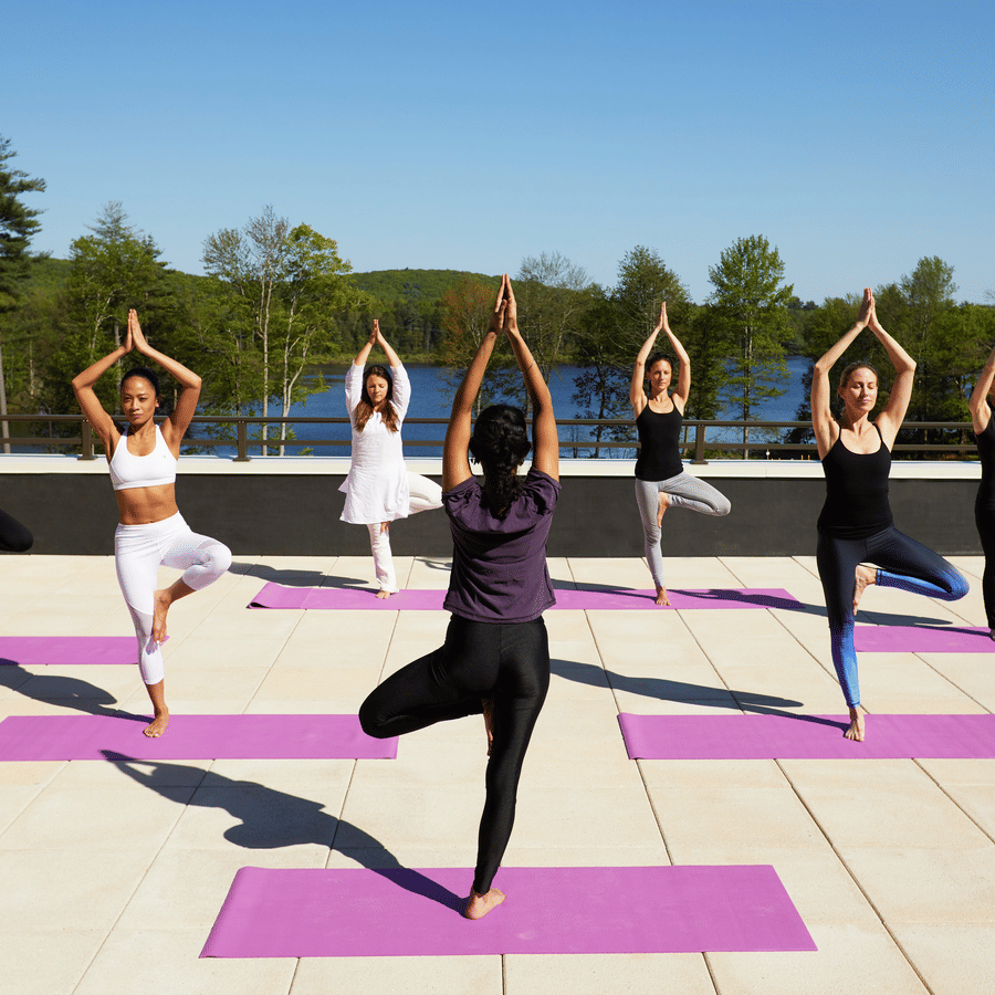 A group of 7 women performing yoga on the rooftop at YO1 Longevity & Health Resorts, Catskills with a backdrop of the lake surrounded by trees