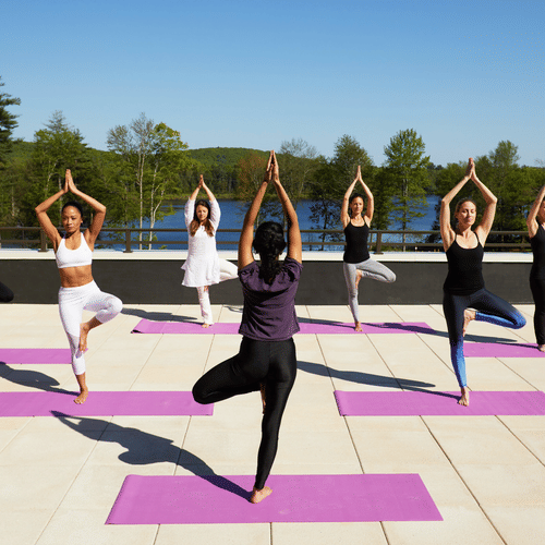 A group of 7 women performing yoga on the rooftop at YO1 Longevity & Health Resorts, Catskills with a backdrop of the lake surrounded by trees
