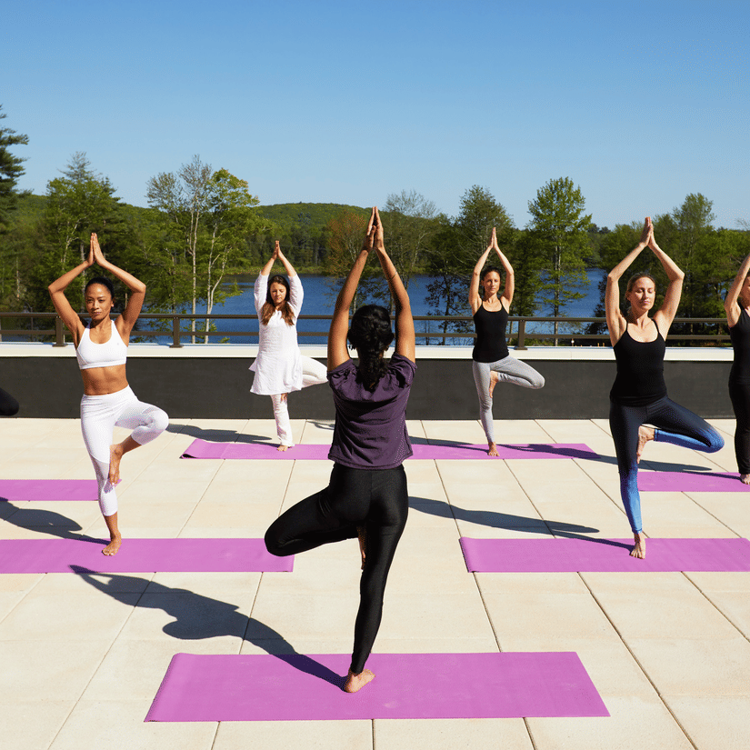 A group of 7 women performing yoga on the rooftop at YO1 Longevity & Health Resorts, Catskills with a backdrop of the lake surrounded by trees